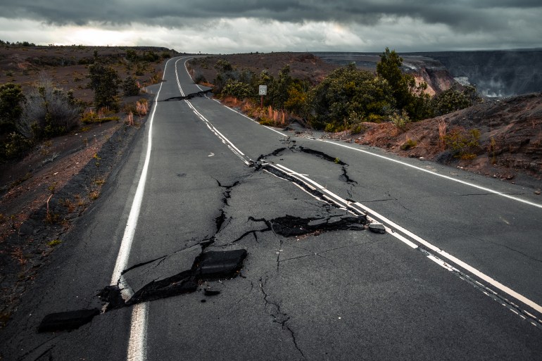 Damaged asphalt road (Crater Rim Drive) in the Hawaii Volcanoes National Park after earthquake and eruption of Kilauea (fume at upper right) volcano in May 2018. Big Island, Hawaii