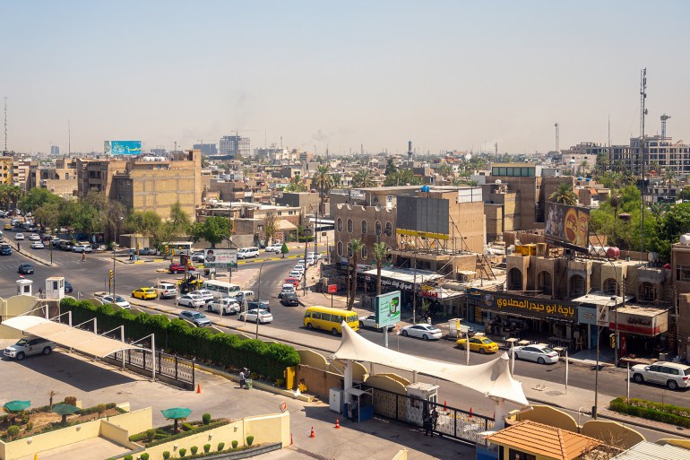 View of a vibrant cityscape of Baghdad, Iraq, with a busy urban scene. Numerous cars, including yellow taxis, fill the streets, and several buildings line the roads with a mix of modern and traditional architecture. The visible shops have signs written in Arabic, with a focus on daily life and commerce. A few palm trees and scattered greenery add contrast to the otherwise dense urban environment. In the background, the skyline stretches out with various structures and taller buildings, while a slightly hazy sky suggests warm and dry weather which is typical of this region. No