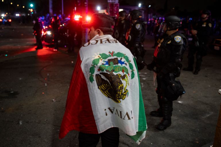 A protester wrapped in a Mexican flag stands next to law enforcement officers as they face off with demonstrators during a protest following federal immigration operations, in the Compton neighborhood of Los Angeles, California early on June 8, 2025.